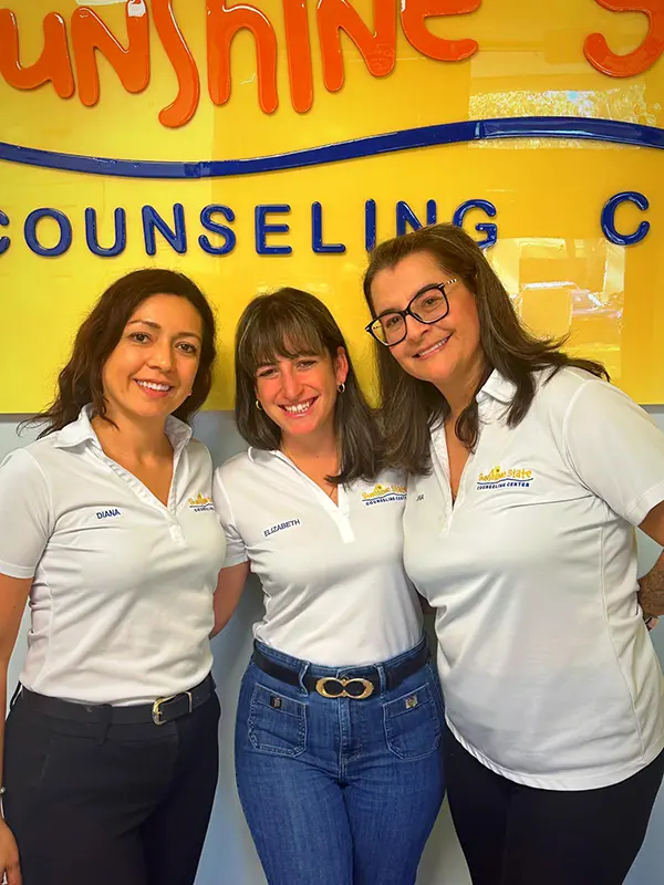 Tres terapeutas sonrientes en camisetas del Sunshine State Counseling Center, fondo amarillo con el logo del centro, representando servicios de salud mental en español.