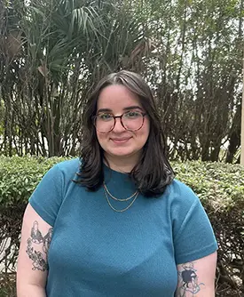 Leinys Torres, consejera en Sunshine State Counseling Center, posando en un entorno natural, con cabello oscuro y gafas, vestida con una camiseta azul, destacando su profesionalismo en servicios de salud mental.